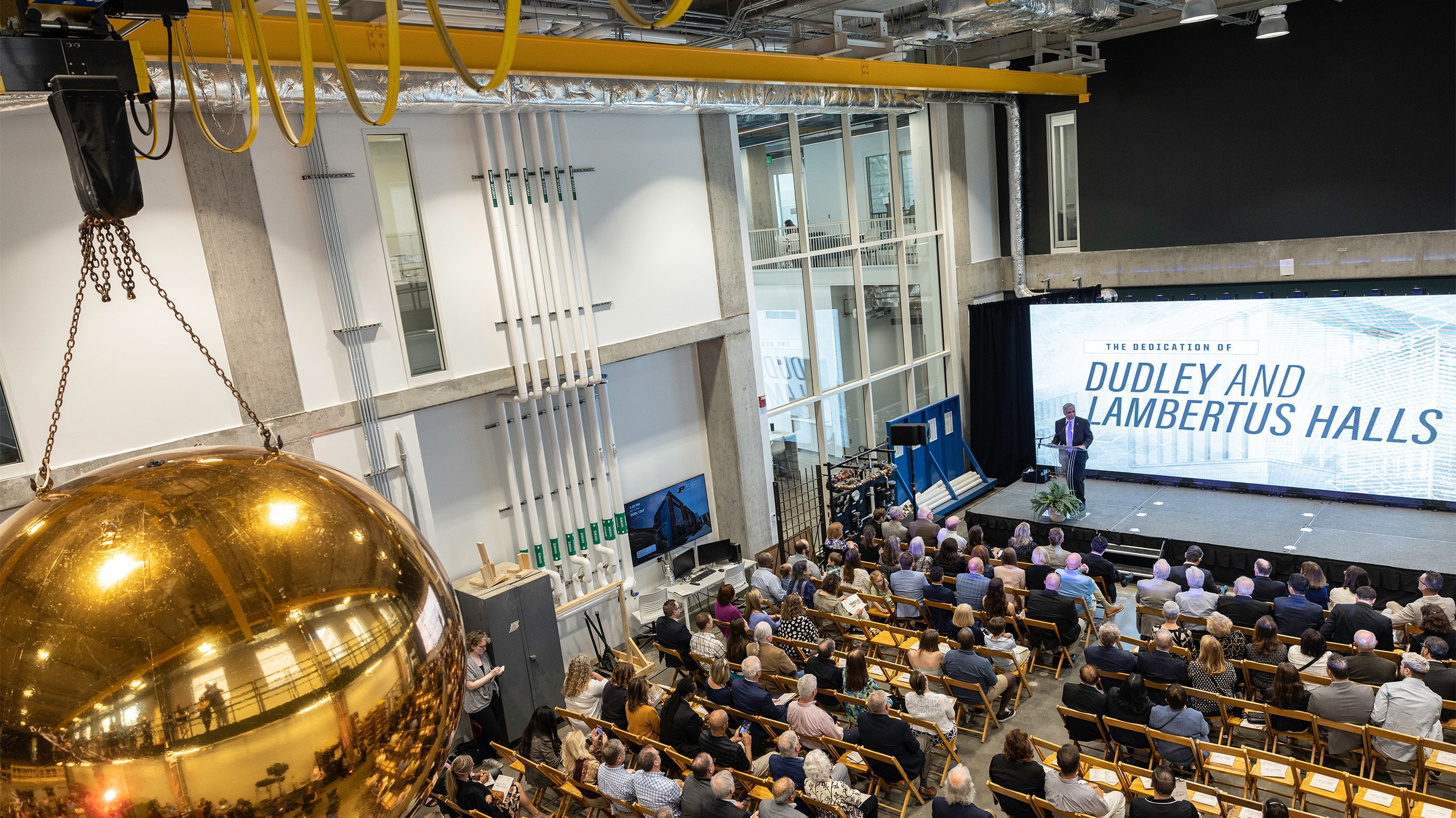 A group of people sitting in front of a stage and a screen that says "The dedication of Dudley and Lambertus Halls"