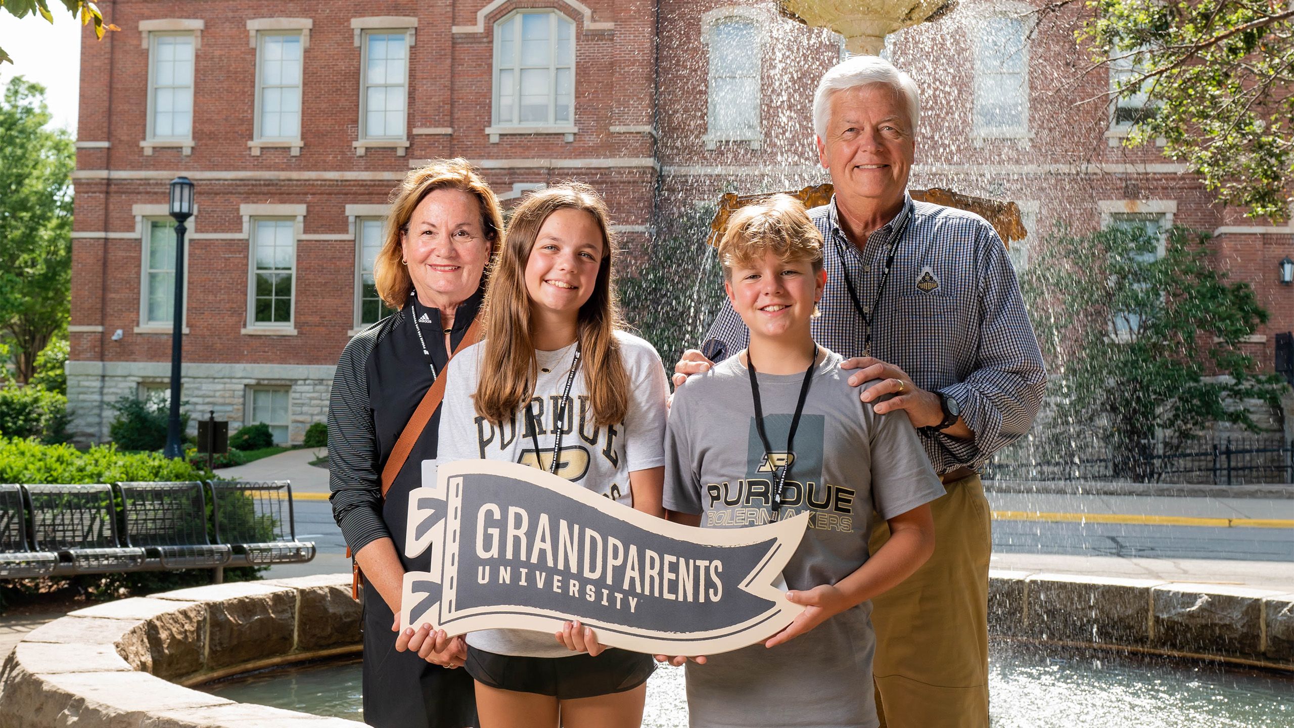 Two grandparents and two grandchildren stand in front of a fountain with a sign that says "Grandparents University"