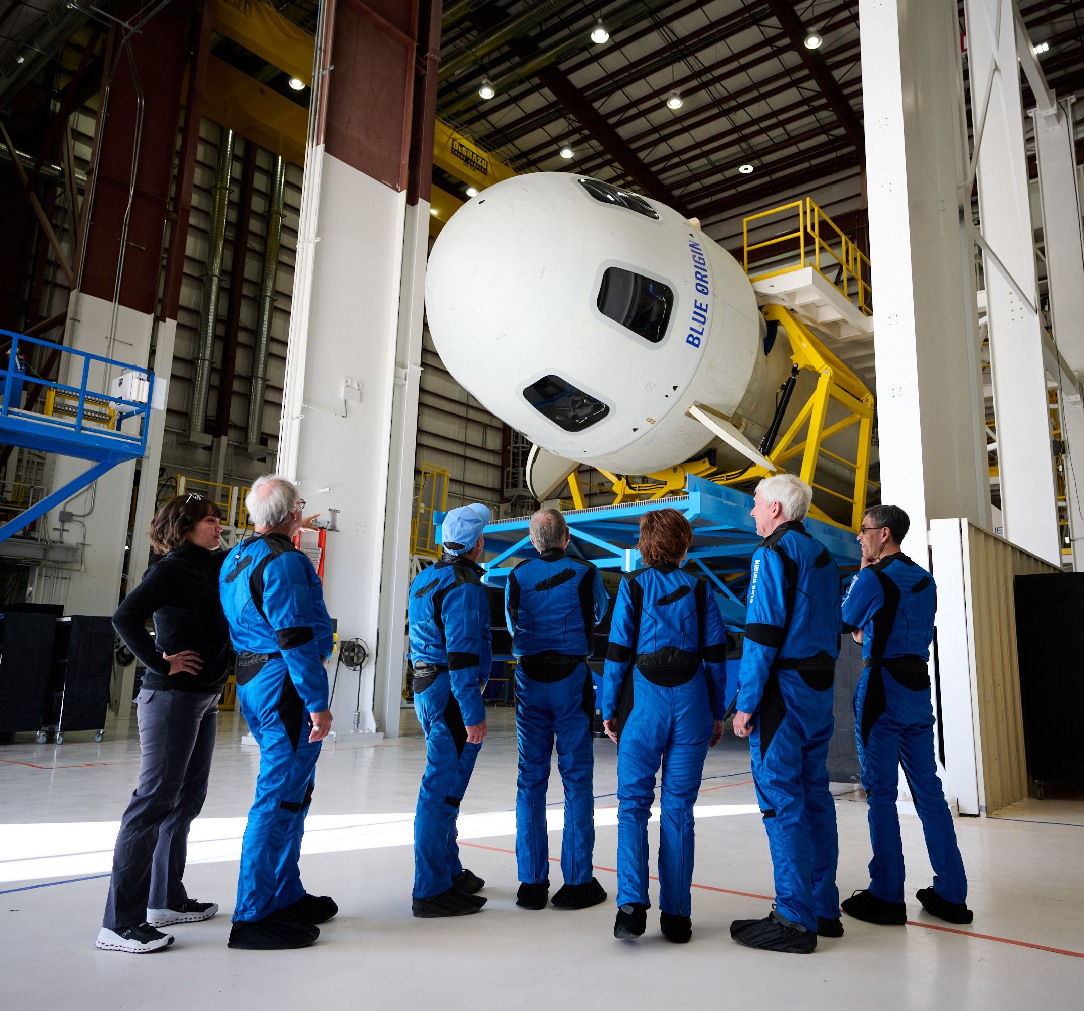 The image shows spacefarers in blue jumpsuits standing in a shuttle hangar