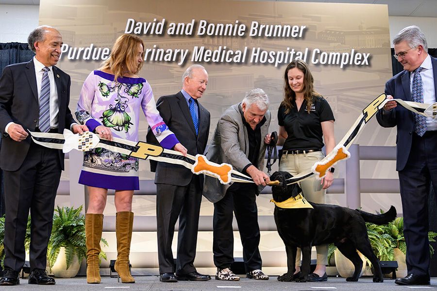 The image shows a group of people and one labrador retriever on stage at a ribbon cutting